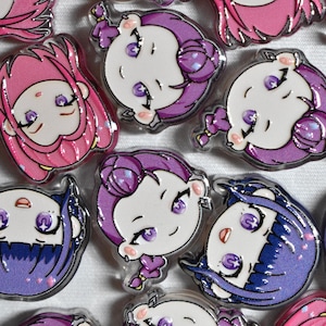 Può includere: Una collezione di piccole spille circolari in acrilico raffiguranti volti di personaggi in stile chibi con grandi occhi viola. I personaggi hanno capelli nelle tonalità del rosa, viola e blu scuro, con sottili riflessi glitterati.