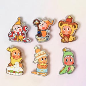 May include: A collection of six colorful cartoon character pins. Each pin features a unique design, including characters in costumes like a bear, a dinosaur, and a chef. The pins have a glossy finish and are set against a white background.