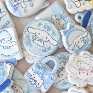 May include: A collection of flat, light blue and white decorative charms featuring the character Cinnamoroll. The charms include various shapes such as rectangles, circles, and a padlock, all decorated with blue bows, floral patterns, and text.