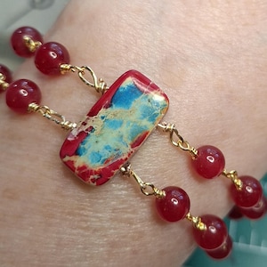 May include: A beaded bracelet featuring a rectangular stone with red, blue, and gold patterns. The bracelet has red beads and gold-colored chain links. The jewelry is designed to be worn on the wrist.