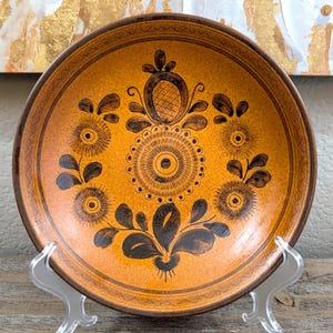 May include: A round, ceramic plate with a warm orange background and intricate black floral designs. The plate has a brown rim and is displayed on a clear plastic stand. The floral pattern includes stylized flowers and leaves.