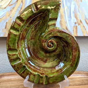 May include: A glossy ceramic ashtray shaped like a spiral seashell, featuring a variegated glaze of moss green and deep brown tones. The curved outer edge includes four distinct notches for resting items, leading into a central spiral design.