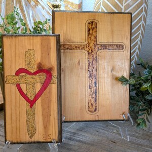 Pode incluir: Duas placas retangulares de madeira com desenhos de cruzes pirografados. A placa menor exibe uma cruz com textura dourada sobreposta por um coração vermelho. A placa maior apresenta uma cruz escura e texturizada. Ambas têm bordas escuras.