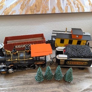 Pode incluir: Um conjunto de comboios modelo com uma locomotiva preta e dourada com um telhado laranja, um vagão de carga vermelho, um vagão amarelo e um vagão de carvão preto. Inclui três pequenas árvores de Natal verdes. O conjunto de comboios está numa superfície de madeira.