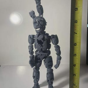 以下が含まれることがあります： 損傷した風化した外観を持つ、ウサギのようなキャラクターのグレーの3Dプリント製可動フィギュア。フィギュアは黄色のメジャーの横に立っており、足から耳までの全長が約25.4cmであることを示しています。