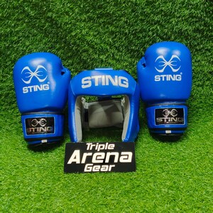 Op de afbeelding: Een set blauwe boksuitrusting bestaande uit twee bokshandschoenen en een hoofdbeschermer, beide voorzien van het witte Sting-logo en tekst. Een zwart rechthoekig bord met de witte tekst Triple Arena Gear staat voor de hoofdbeschermer.