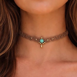 Può includere: Una collana girocollo marrone con un pendente turchese. La collana è realizzata in materiale intrecciato e presenta piccole perline color oro. Il pendente ha la forma di una goccia. La modella indossa un top color crema con spalle scoperte.