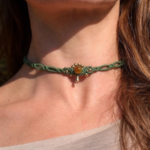 May include: A green macrame choker necklace with a central floral design. The necklace features a small, round, amber-colored stone and gold-colored beads. A small gold-colored pendant hangs below the central design.