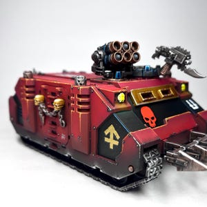 Puede incluir: Modelo detallado de un vehículo blindado rojo y negro con un emblema de calavera y múltiples sistemas de armas. El tanque en miniatura presenta un lanzacohetes múltiple, una cuchilla y un emblema de calavera. El modelo está pintado con detalles dorados y tiene una base con orugas.