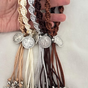 K&ouml;nnte beinhalten: Eine Sammlung von Makramee-Armb&auml;ndern in verschiedenen neutralen Farben, darunter Beige, Creme, Grau, Braun und Dunkelbraun. Jedes Armband hat eine silberfarbene Medaille und baumelnde Anh&auml;nger. Die Armb&auml;nder sind vor wei&szlig;em Hintergrund ausgestellt.