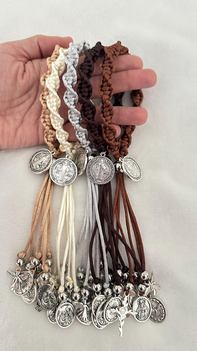 K&ouml;nnte beinhalten: Eine Sammlung von Makramee-Armb&auml;ndern in verschiedenen neutralen Farben, darunter Beige, Creme, Grau, Braun und Dunkelbraun. Jedes Armband hat eine silberfarbene Medaille und baumelnde Anh&auml;nger. Die Armb&auml;nder sind vor wei&szlig;em Hintergrund ausgestellt.