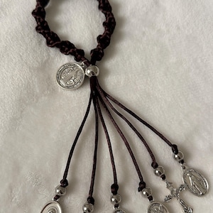 K&ouml;nnte beinhalten: Ein dunkelbraunes, geknotetes Armband mit baumelnden Anh&auml;ngern. Die Anh&auml;nger umfassen silberfarbene religi&ouml;se Medaillen, ein Kreuz und einen Engel. Das Armband wird auf einer wei&szlig;en, strukturierten Oberfl&auml;che pr&auml;sentiert.