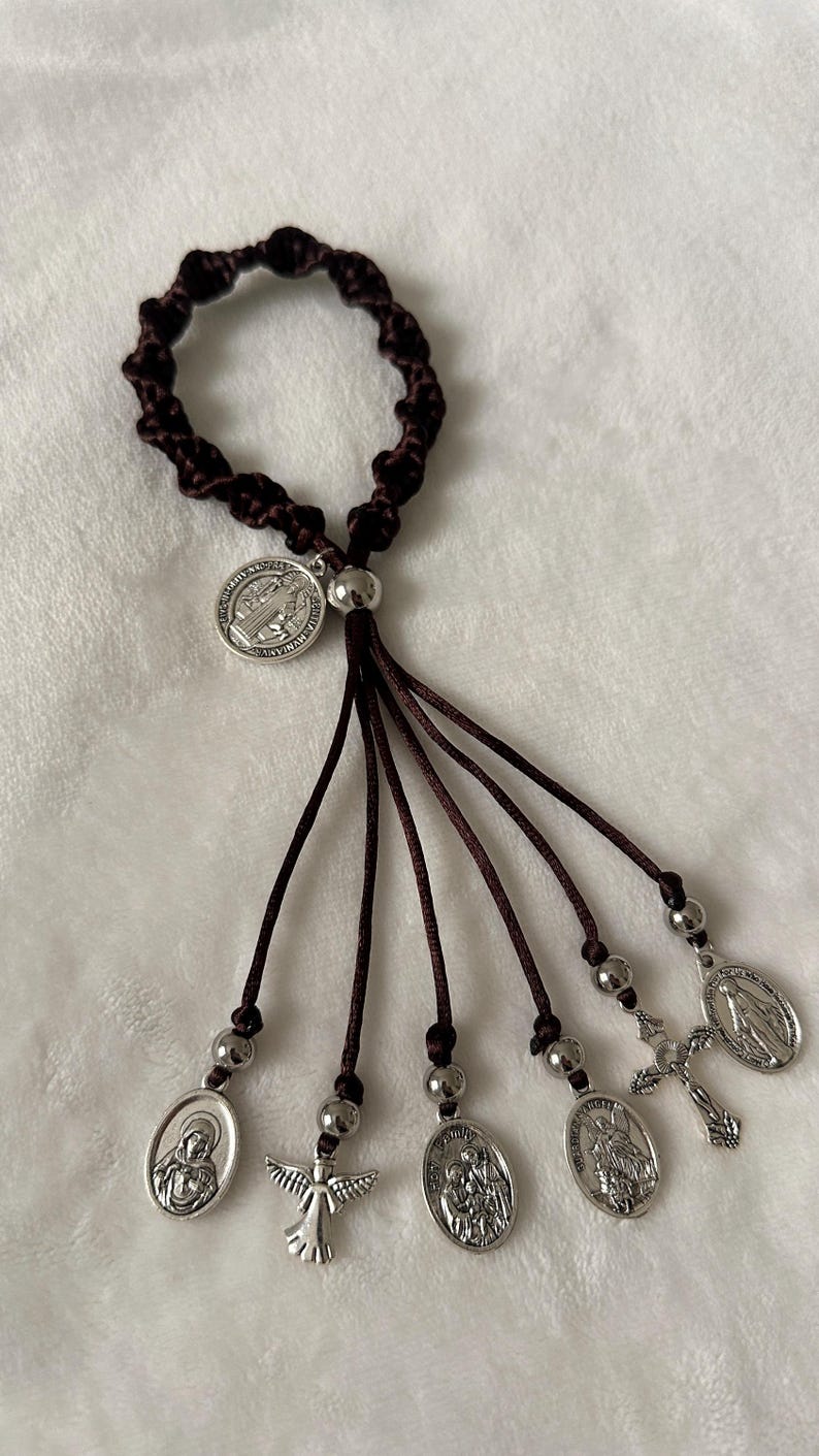 K&ouml;nnte beinhalten: Ein dunkelbraunes, geknotetes Armband mit baumelnden Anh&auml;ngern. Die Anh&auml;nger umfassen silberfarbene religi&ouml;se Medaillen, ein Kreuz und einen Engel. Das Armband wird auf einer wei&szlig;en, strukturierten Oberfl&auml;che pr&auml;sentiert.