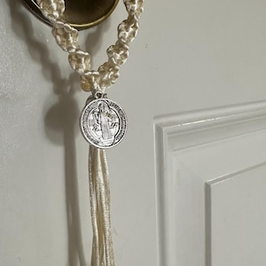 K&ouml;nnte beinhalten: Cremefarbener Makramee-Rosenkranz, der an einem Messing-T&uuml;rknauf h&auml;ngt. Der Rosenkranz hat eine silberne St. Benedikt-Medaille und mehrere kleinere silberne Medaillons. Die Schnur ist in einem dekorativen Muster geknotet.