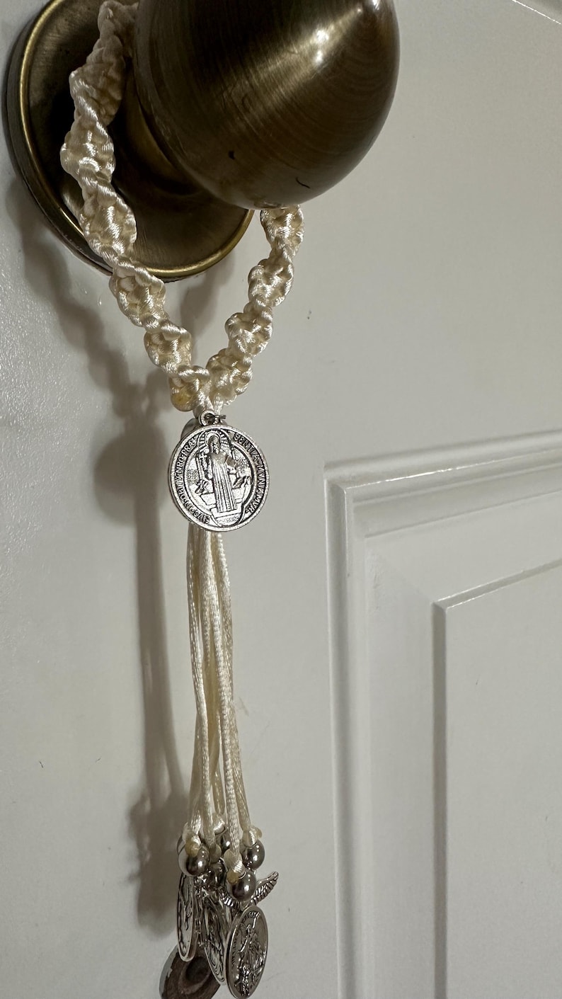 K&ouml;nnte beinhalten: Cremefarbener Makramee-Rosenkranz, der an einem Messing-T&uuml;rknauf h&auml;ngt. Der Rosenkranz hat eine silberne St. Benedikt-Medaille und mehrere kleinere silberne Medaillons. Die Schnur ist in einem dekorativen Muster geknotet.