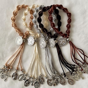 K&ouml;nnte beinhalten: Eine Sammlung von Rosenkranz-Armb&auml;ndern in verschiedenen Farben, darunter Beige, Creme, Silber, Braun und Burgunder. Jedes Armband hat ein geflochtenes Design, eine silberne Medaille und eine Quaste aus kleinen Charms und Perlen.
