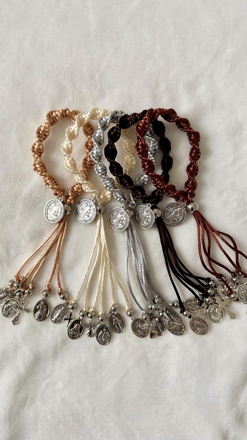 K&ouml;nnte beinhalten: Eine Sammlung von Rosenkranz-Armb&auml;ndern in verschiedenen Farben, darunter Beige, Creme, Silber, Braun und Burgunder. Jedes Armband hat ein geflochtenes Design, eine silberne Medaille und eine Quaste aus kleinen Charms und Perlen.