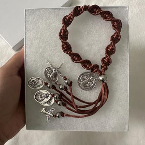 K&ouml;nnte beinhalten: Ein brauner Makramee-Rosenkranz mit silberfarbenen Metallakzenten, einschlie&szlig;lich Medaillons und Perlen. Der Rosenkranz wird in einer wei&szlig;en Geschenkbox pr&auml;sentiert, die sein aufwendiges Design und seine religi&ouml;se Bedeutung hervorhebt.