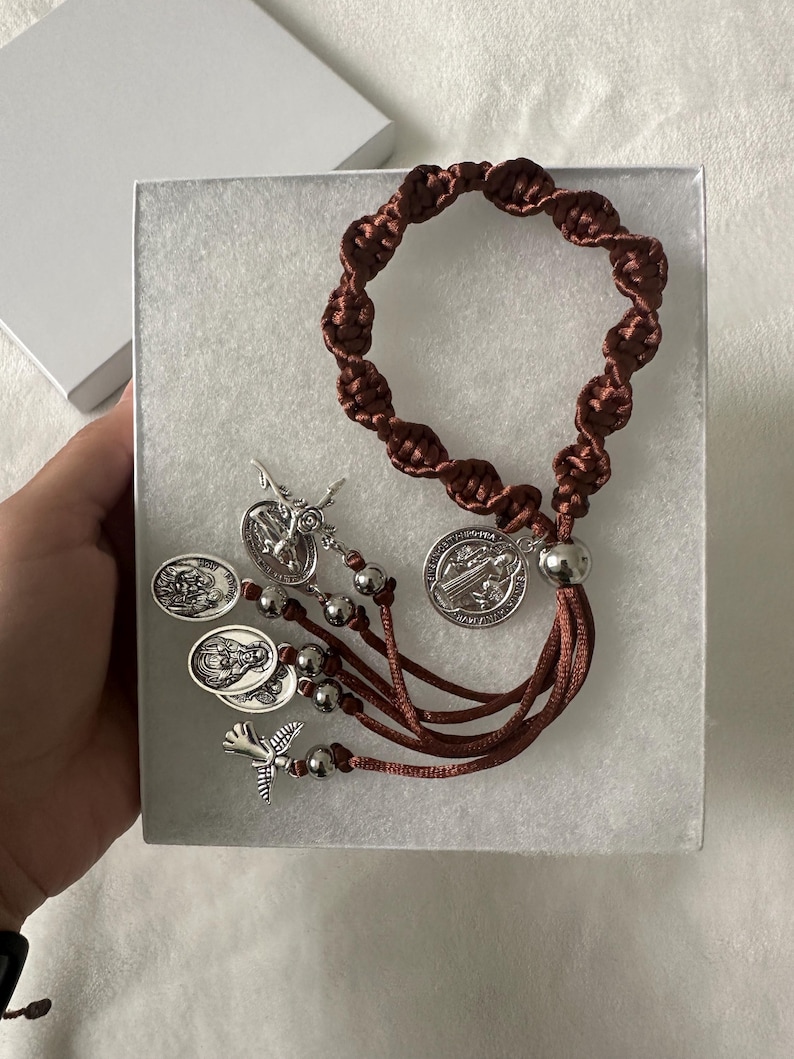 K&ouml;nnte beinhalten: Ein brauner Makramee-Rosenkranz mit silberfarbenen Metallakzenten, einschlie&szlig;lich Medaillons und Perlen. Der Rosenkranz wird in einer wei&szlig;en Geschenkbox pr&auml;sentiert, die sein aufwendiges Design und seine religi&ouml;se Bedeutung hervorhebt.
