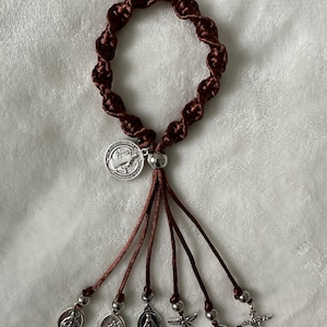 K&ouml;nnte beinhalten: Ein braunes Rosenkranzarmband mit geflochtenem Design. Das Armband verf&uuml;gt &uuml;ber ein silberfarbenes Medaillon und mehrere baumelnde Anh&auml;nger, darunter Heiligenmedaillen und ein kleines Kreuz. Der Rosenkranz wird auf einer wei&szlig;en, strukturierten Oberfl&auml;che pr&auml;sentiert.