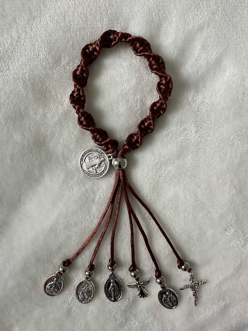 K&ouml;nnte beinhalten: Ein braunes Rosenkranzarmband mit geflochtenem Design. Das Armband verf&uuml;gt &uuml;ber ein silberfarbenes Medaillon und mehrere baumelnde Anh&auml;nger, darunter Heiligenmedaillen und ein kleines Kreuz. Der Rosenkranz wird auf einer wei&szlig;en, strukturierten Oberfl&auml;che pr&auml;sentiert.