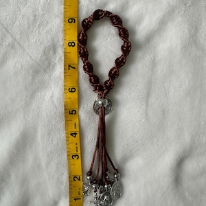 K&ouml;nnte beinhalten: Ein brauner Makramee-Rosenkranz mit silberfarbenen Anh&auml;ngern und einem Kreuzanh&auml;nger. Der Rosenkranz ist etwa 20 cm lang, gemessen mit einem gelben Ma&szlig;band. Die Anh&auml;nger umfassen religi&ouml;se Figuren und einen kleinen Engel.