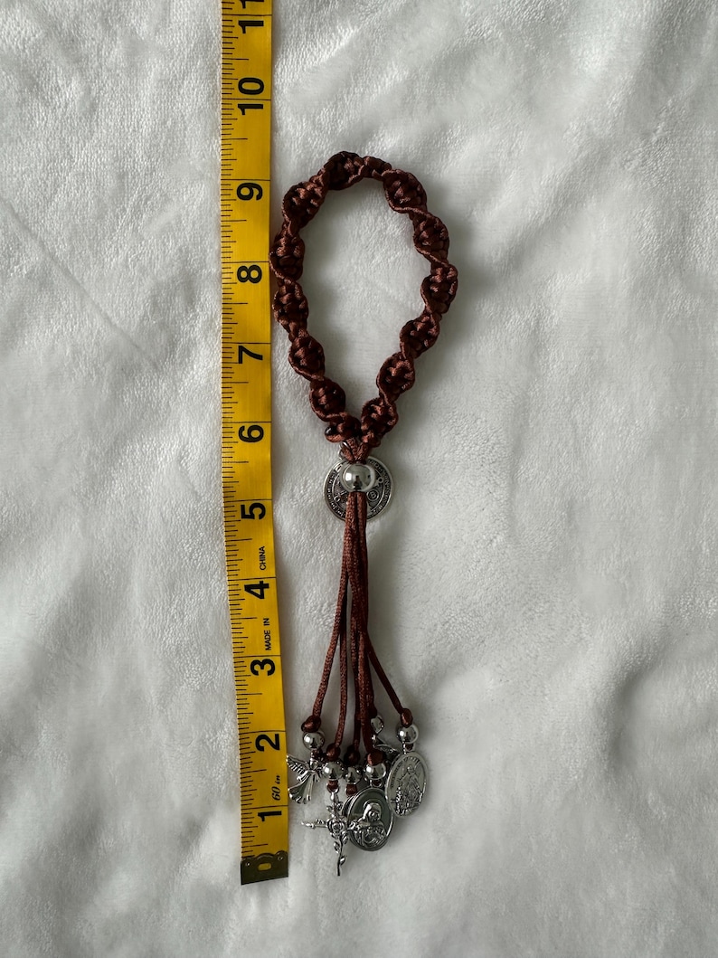 K&ouml;nnte beinhalten: Ein brauner Makramee-Rosenkranz mit silberfarbenen Anh&auml;ngern und einem Kreuzanh&auml;nger. Der Rosenkranz ist etwa 20 cm lang, gemessen mit einem gelben Ma&szlig;band. Die Anh&auml;nger umfassen religi&ouml;se Figuren und einen kleinen Engel.