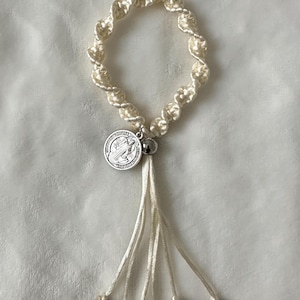 K&ouml;nnte beinhalten: Cremefarbenes Makramee-Armband mit silbernen Anh&auml;ngern und Charms. Das Armband hat ein kreisf&ouml;rmiges Design mit einer Quaste aus Kordeln. Zu den Charms geh&ouml;ren ein Kreuz, ein Engel und religi&ouml;se Medaillons. Das Armband ist vor einem wei&szlig;en Hintergrund platziert.