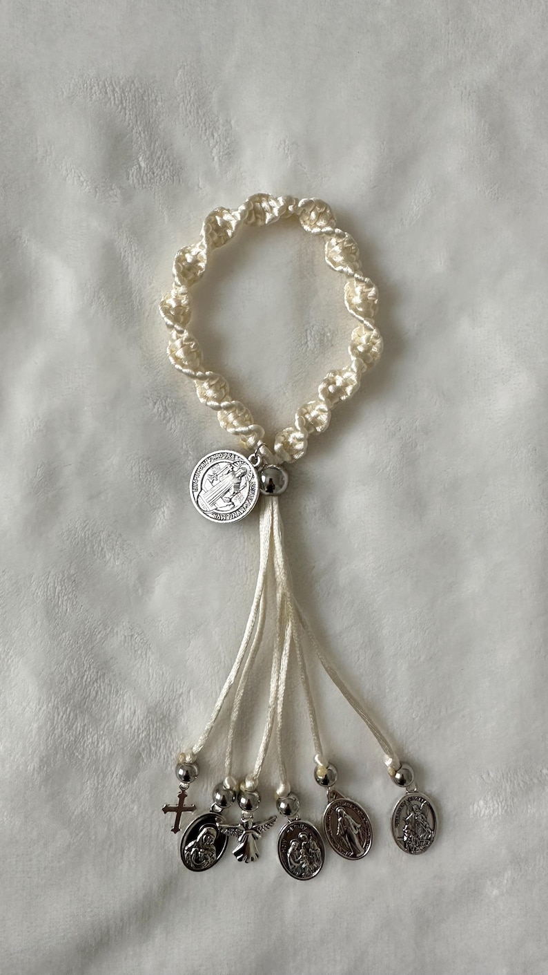 K&ouml;nnte beinhalten: Cremefarbenes Makramee-Armband mit silbernen Anh&auml;ngern und Charms. Das Armband hat ein kreisf&ouml;rmiges Design mit einer Quaste aus Kordeln. Zu den Charms geh&ouml;ren ein Kreuz, ein Engel und religi&ouml;se Medaillons. Das Armband ist vor einem wei&szlig;en Hintergrund platziert.
