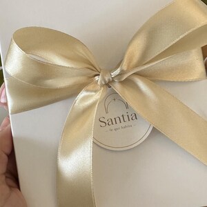 K&ouml;nnte beinhalten: Eine wei&szlig;e Geschenkbox, gebunden mit einem goldenen Satinband und einer Schleife. Die Box hat ein rundes Etikett mit dem Wort "Santia" und dem Satz "fe que habita". Rosa und rote Tulpen sind im Hintergrund.