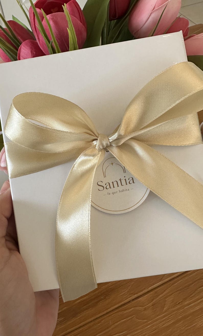 K&ouml;nnte beinhalten: Eine wei&szlig;e Geschenkbox, gebunden mit einem goldenen Satinband und einer Schleife. Die Box hat ein rundes Etikett mit dem Wort "Santia" und dem Satz "fe que habita". Rosa und rote Tulpen sind im Hintergrund.