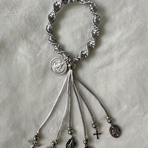 K&ouml;nnte beinhalten: Ein silbernes und graues Rosenkranzarmband mit geflochtenem Design. Das Armband verf&uuml;gt &uuml;ber ein zentrales Medaillon und mehrere baumelnde Charms, darunter ein Engel, ein Kreuz und andere religi&ouml;se Symbole. Das Armband wird auf einer weichen, wei&szlig;en Oberfl&auml;che pr&auml;sentiert.