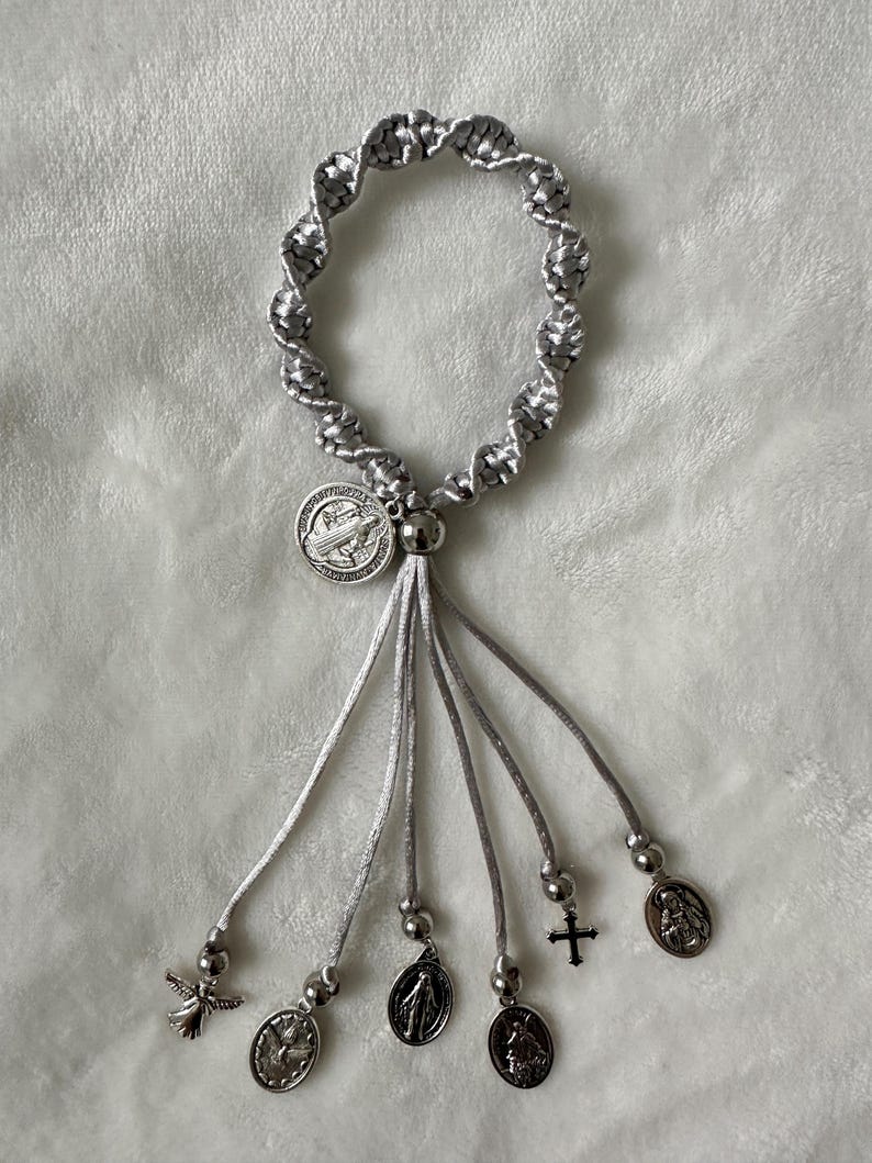 K&ouml;nnte beinhalten: Ein silbernes und graues Rosenkranzarmband mit geflochtenem Design. Das Armband verf&uuml;gt &uuml;ber ein zentrales Medaillon und mehrere baumelnde Charms, darunter ein Engel, ein Kreuz und andere religi&ouml;se Symbole. Das Armband wird auf einer weichen, wei&szlig;en Oberfl&auml;che pr&auml;sentiert.