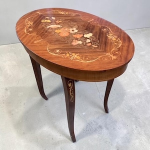 May include: An oval wooden side table featuring a rich brown finish and intricate floral marquetry inlay on the tabletop. The table stands on four curved legs, each decorated with a delicate light-coloured vine inlay pattern along the front surface.