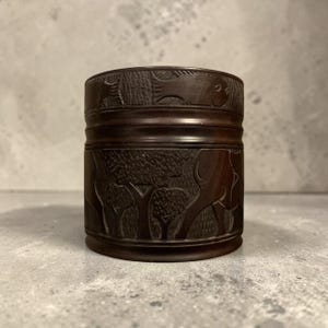 May include: A dark brown, cylindrical wooden container with detailed carvings. The container features relief carvings of elephants, trees, and other wildlife. The design wraps around the entire container, showcasing a natural theme.