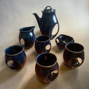 Puede incluir: Un juego de té de cerámica que incluye una tetera alta con tapa, una jarra para crema y cuatro tazas. Cada pieza tiene un acabado en esmalte azul marino brillante y se caracteriza por un diseño de corte circular integrado en la base.