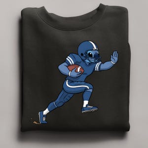 May include: Black sweatshirt with a cartoon character in a blue American football uniform, helmet, and holding a football. The character is running with one arm raised. The design includes white stripes and accents.