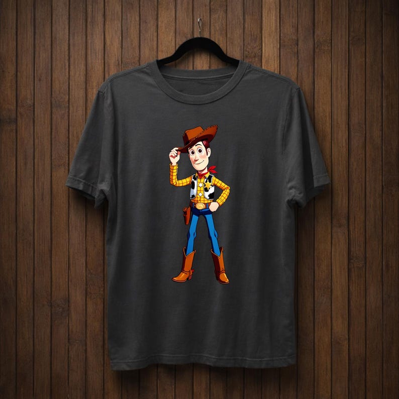 May include: Dark grey t-shirt with a cartoon cowboy character illustration. The cowboy wears a brown hat, yellow vest, blue jeans, and brown boots. The character is tipping his hat, with a red neckerchief.