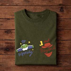 Op de afbeelding: Olijfgroen t-shirt met een cartoonontwerp. Het ontwerp bevat een ruimteschip en een cowboyhoed, met de woorden "to infinity and beyond" in blauw, rood en geel.