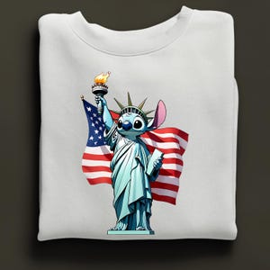 Puede incluir: Sudadera blanca con un personaje de dibujos animados como la Estatua de la Libertad, sosteniendo una antorcha y una tableta, con una bandera estadounidense de fondo. El personaje es azul con ojos y orejas grandes. Dise&ntilde;o patri&oacute;tico y caprichoso.