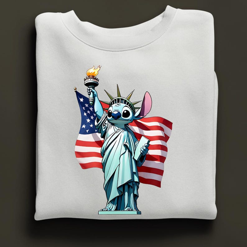 Puede incluir: Sudadera blanca con un personaje de dibujos animados como la Estatua de la Libertad, sosteniendo una antorcha y una tableta, con una bandera estadounidense de fondo. El personaje es azul con ojos y orejas grandes. Dise&ntilde;o patri&oacute;tico y caprichoso.