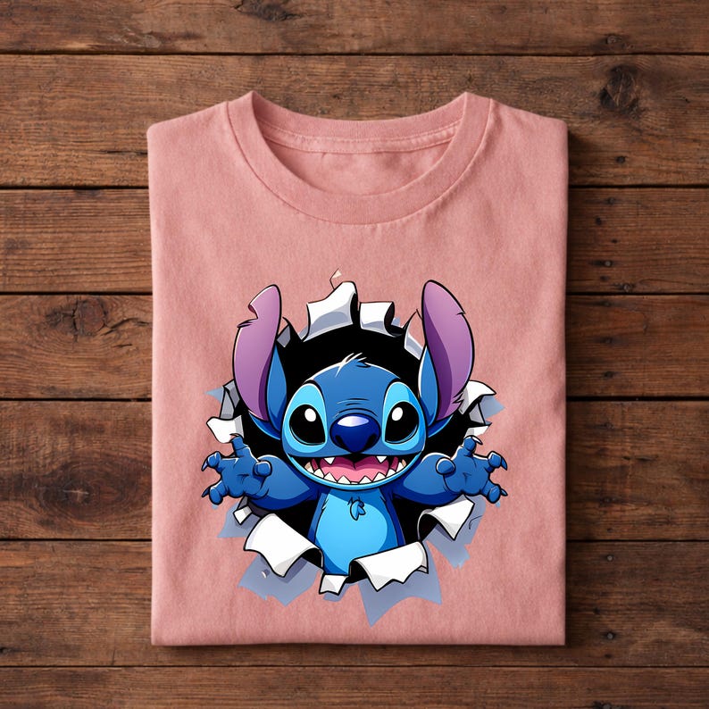 May include: A light pink t-shirt featuring a cartoon graphic of Stitch bursting through a torn hole. The blue character has large eyes, a wide grin, and raised arms. The design is centered on the folded shirt.