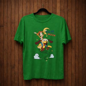 Peut inclure: T-shirt vert avec un dessin anim&eacute; d'un personnage de cow-boy chevauchant un chien, avec le texte "Fly me to the Moon". Le motif comprend des &eacute;toiles, des nuages et un croissant de lune.