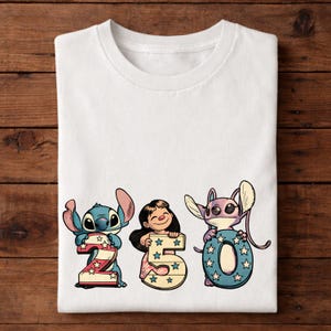 Puede incluir: Camiseta blanca con los personajes Stitch y Lilo sosteniendo los n&uacute;meros 2, 5 y 0. Los n&uacute;meros est&aacute;n decorados con estrellas y colores patri&oacute;ticos. La camiseta est&aacute; doblada sobre una superficie de madera.