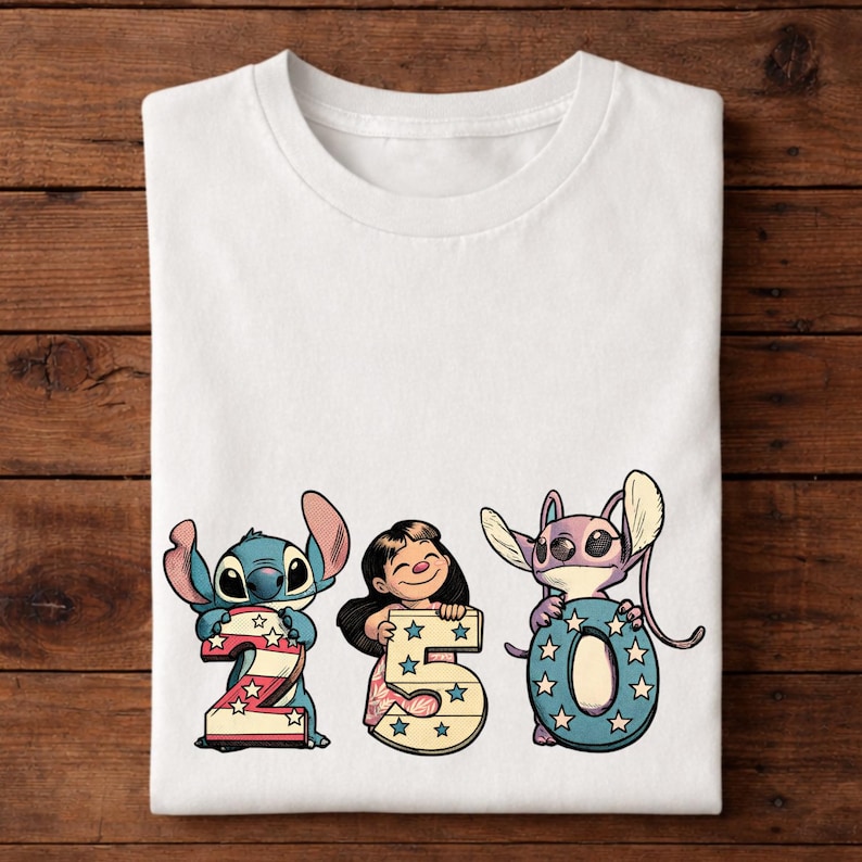 Puede incluir: Camiseta blanca con los personajes Stitch y Lilo sosteniendo los n&uacute;meros 2, 5 y 0. Los n&uacute;meros est&aacute;n decorados con estrellas y colores patri&oacute;ticos. La camiseta est&aacute; doblada sobre una superficie de madera.