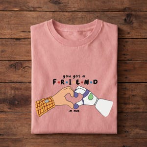 May include: A pink t-shirt with the text "you got a FRIEND in me" and a graphic of two hands forming a heart. One hand is a cartoon character's arm with a plaid cuff, and the other is a robot arm.