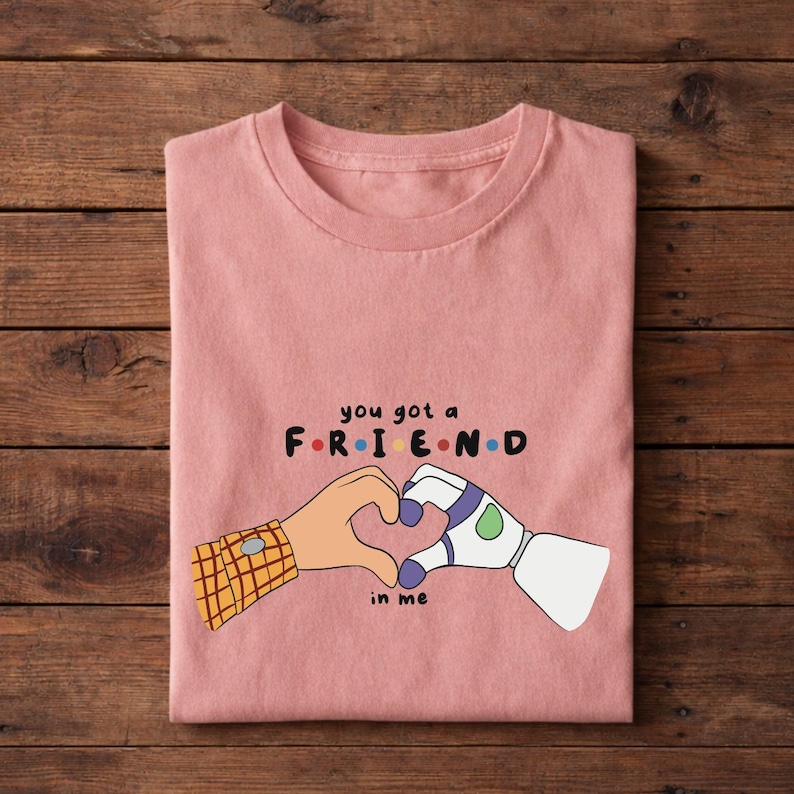 May include: A pink t-shirt with the text "you got a FRIEND in me" and a graphic of two hands forming a heart. One hand is a cartoon character's arm with a plaid cuff, and the other is a robot arm.