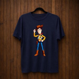 May include: Navy blue t-shirt with a cartoon cowboy character illustration. The cowboy wears a brown hat, yellow vest, red neckerchief, blue trousers, and brown boots. The character is tipping their hat.