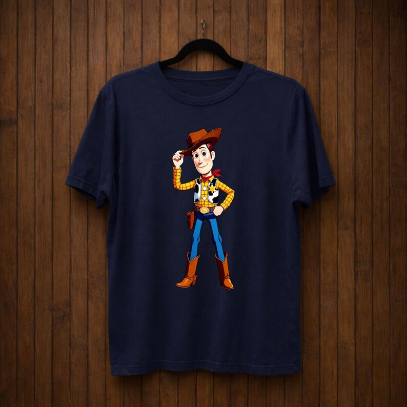 May include: Navy blue t-shirt with a cartoon cowboy character illustration. The cowboy wears a brown hat, yellow vest, red neckerchief, blue trousers, and brown boots. The character is tipping their hat.