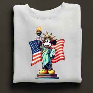 Puede incluir: Sudadera blanca con una representaci&oacute;n de dibujos animados de Mickey Mouse como la Estatua de la Libertad, sosteniendo una antorcha y una tableta, con una bandera estadounidense de fondo. El dise&ntilde;o es colorido y patri&oacute;tico.